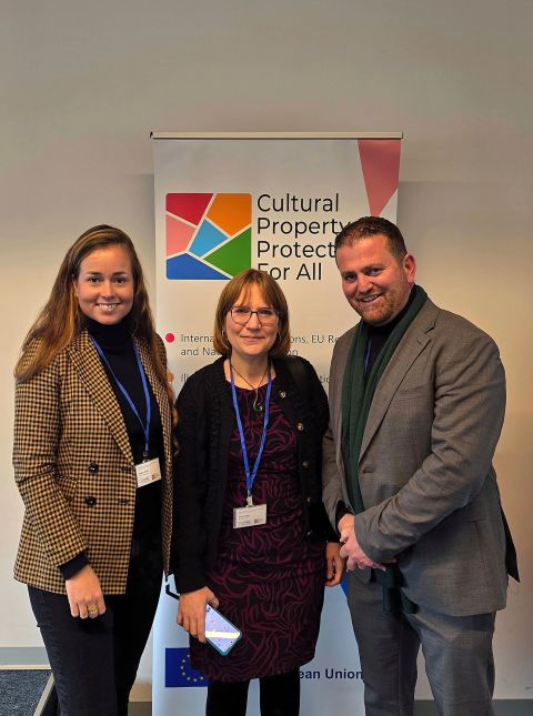 Three people standing in front of a standing banner that says Cultural Property Protection for All. Two women and one man. They are wearing conference name tags and are smiling.
