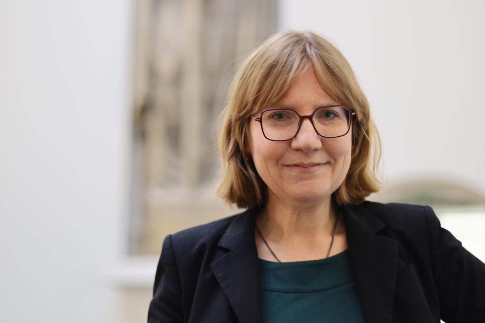 photograph of donna yates, a white woman in her 40s with red hair, glasses, and wearing a black blazer in front of a blurry statue.